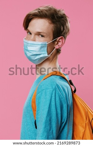 portrait of a handsome peaceful red-haired student man with a backpack in a medical mask on his face looking at the camera. Horizontal photo on a pink background with empty space for advertising text