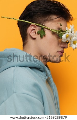a close portrait of a handsome, funny man with daisies on his eyes, standing on a bright background in a light blue hoodie with his head turned to the side