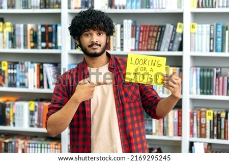 Handsome curly mixed race guy, in casual clothes, stands in the library against the background of bookshelves, holds a sign in his hand that says looking for a job, looks at camera, hopes to get a job Royalty-Free Stock Photo #2169295245