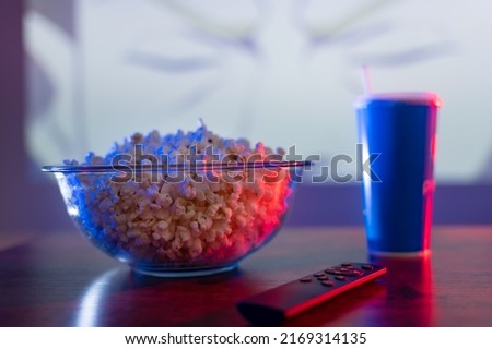 Close-up. On a wooden table are a glass bowl of popcorn, a carbonated drink in a plastic glass, and a TV remote control. Home comfort, watching your favorite movies, TV shows, relaxing.