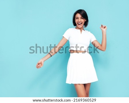 Young beautiful smiling female in trendy summer white top and skirt. Carefree woman posing near blue wall in studio. Positive model having fun indoors. Cheerful and happy. Celebrating and dancing  Royalty-Free Stock Photo #2169360529