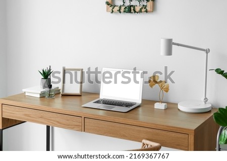 Modern workplace with blank frame and laptop near light wall