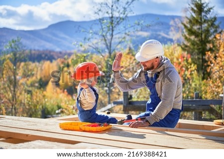 Father with toddler son building wooden frame house. Male builder giving high five to kid on construction site, wearing helmet and blue overalls on sunny day. Carpentry and family concept. Royalty-Free Stock Photo #2169388421