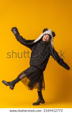 Portrait of a cheerful cute teenage girl with long black hair, dressed in a panda fur hat, posing on a yellow studio background. comical image. Advertisement. original children's clothing. full growth