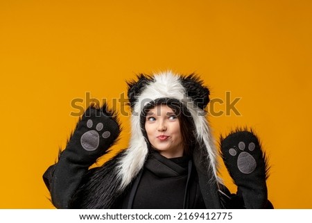 Portrait of a cheerful cute teenage girl with long black hair, dressed in a panda fur hat, posing on a yellow studio background. A comical image. Advertisement. original children's clothing. Close-up