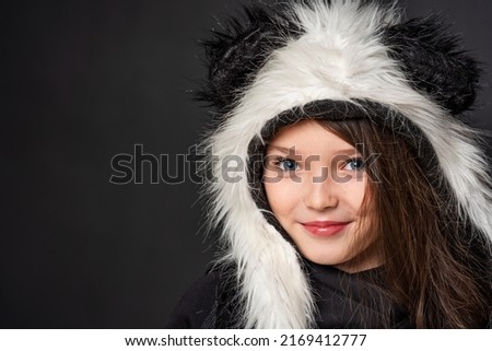 Portrait of a cheerful cute teenage girl with long black hair, dressed in a panda fur hat, posing on a black studio background. A comical image. Advertisement. original children's clothing. Close-up