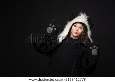A cheerful cute teenage girl with long black hair, dressed in a panda fur hat, poses on a yellow studio background. A comical image. Advertisement
