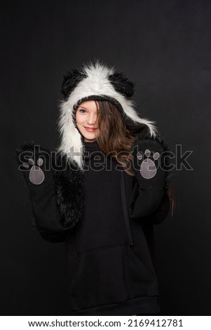 Portrait of a cheerful cute teenage girl with long black hair, dressed in a panda fur hat, posing on a black studio background. A comical image. Advertisement. original children's clothing.