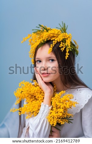 Portrait of cute little girl with long hair and a white dress and a wreath of flowers on her head. child is sitting on chair and holding a bouquet yellow mimosa flowers on blue background in studio