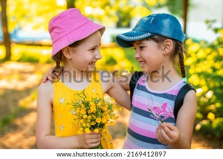 Two funny little girls of different races, in dresses and baseball caps, with a bouquet of wildflowers, laughing and hugging. Best friends like to chat and walk in the fresh air. Summer holidays Royalty-Free Stock Photo #2169412897