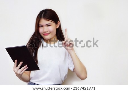 The young asian woman wear white t-shirt is smiling while holding a tablet and pointing up.