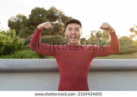 Muscular Asian man flexing biceps with both arms outdoors at sunset Royalty-Free Stock Photo #2169434605