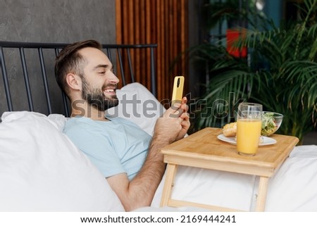 Side view young man in blue t-shirt lying in bed have breakfast take picture shot on mobile cell phone of food rest relax spend time in bedroom lounge home in own room house. Good mood bedtime concept