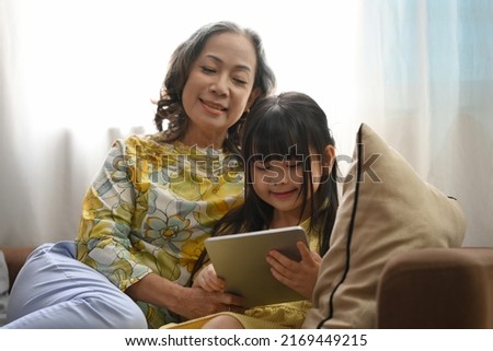 Smiling mature grandma and little preschooler granddaughter using digital tablet surfing internet, watching online cartoon together on couch at home