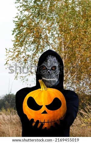 Close-up portrait of grim reaper. Man in death mask with fire flame in eyes on nature yellow birch tree background. Carnival costume, creepy teeth. Halloween holiday concept. Dark horror.