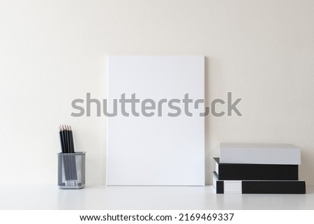 Front view of modern workspace with blank canvas frame on white desk and white wall background, with books and office stationery. Workplace for presentation and mockup.
