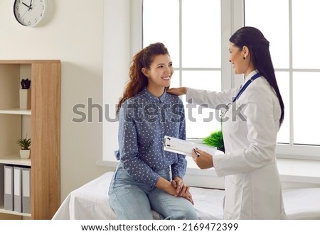 Friendly smiling female doctor touches patient's shoulder and tells her good news. Joyful young woman sitting on couch in doctor's office during medical examination and talking to doctor. Royalty-Free Stock Photo #2169472399
