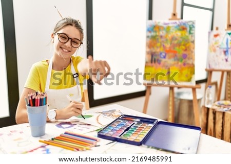 Young brunette teenager at art studio pointing to you and the camera with fingers, smiling positive and cheerful 