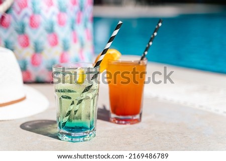 Tropical sparkling lemonade cocktails by the pool with pink beach bag and white hat in the background. Picture of glasses with orange and mint lemon fruit cocktails. Hello summer holiday vacation.