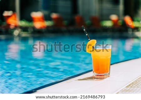 Tropical sparkling cocktail by the pool. The picture of glass with orange lemonade fruit cocktail standing near the poolside. Summer alcohol free drink by the hotel pool. Hello summer holiday vacation Royalty-Free Stock Photo #2169486793