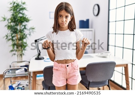 Young hispanic girl standing at pediatrician clinic pointing down looking sad and upset, indicating direction with fingers, unhappy and depressed. 