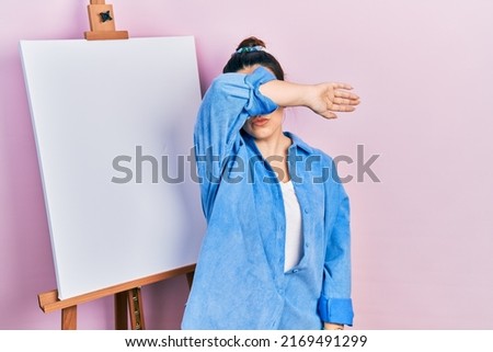 Young hispanic woman standing by painter easel stand covering eyes with arm, looking serious and sad. sightless, hiding and rejection concept 
