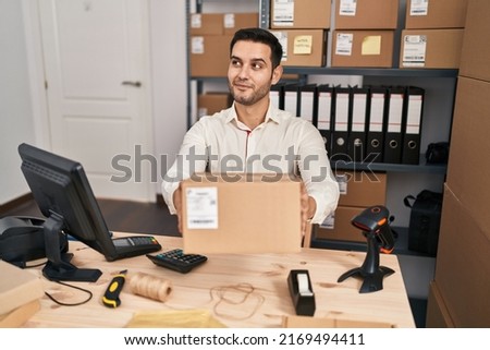 Young hispanic man with beard working at small business ecommerce holding box smiling looking to the side and staring away thinking. 