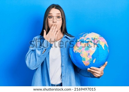 Young hispanic girl holding world ball covering mouth with hand, shocked and afraid for mistake. surprised expression 