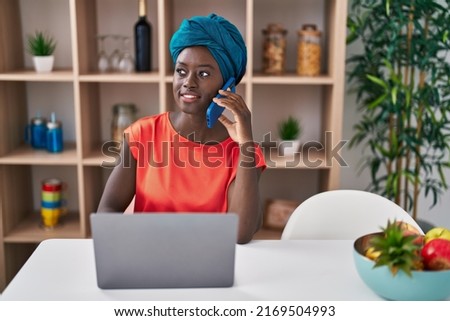Young african american woman talking on the smartphone using laptop at home