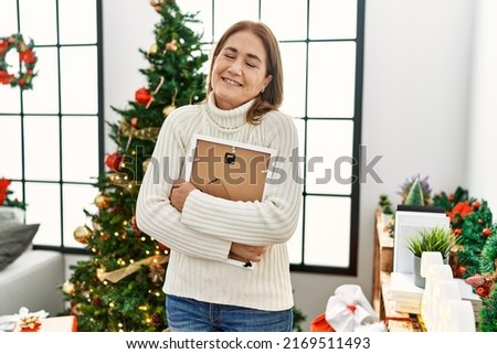 Middle age caucasian woman hugging picture standing by christmas tree at home