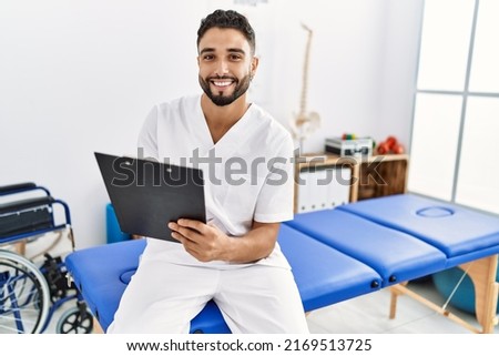 Young arab man wearing physiotherapist uniform writing on clipboard at clinic