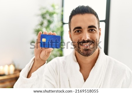 Young hispanic man wearing bathrobe holding credit card at beauty center