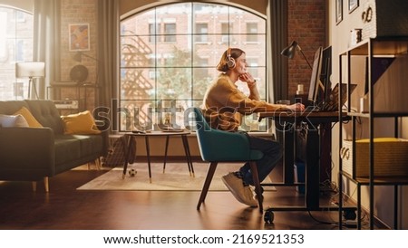 Young Handsome Man Working from Home on Desktop Computer in Sunny Stylish Loft Apartment. Creative Designer Wearing Cozy Yellow Sweater and Headphones. Urban City View from Big Window. Royalty-Free Stock Photo #2169521353