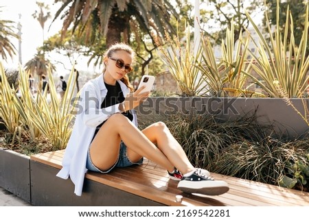 Full-lenght photo of stylish pretty girl wearing blue shirt and shorts in sunglasses is siting on street bench and using smartphone in sunlight