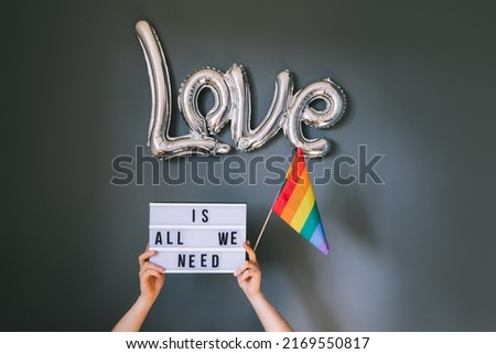 LGBTQ pride month background. Male hands holding lightbox with Text Love is all we need under silver foil balloon with rainbow flag on gray wall background. Film effect tonning. Selective focus.