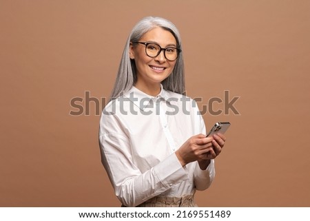Happy business woman in formal shirt touching to the smartphone screen while feeling great with new phone features. Indoor studio shot isolated on beige background Royalty-Free Stock Photo #2169551489