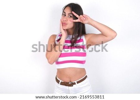 Young beautiful brunette woman wearing striped crop top over white wall  making v-sign near eyes. Leisure lifestyle people person celebrate flirt coquettish concept.