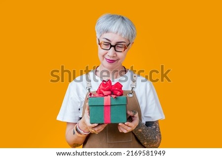 old senior asian female woman with glasses and tattoo wear brown apron hand holding present gift box to camera,smiling cheerful elder woman prepare present green box red ribbon isolate on yellow color Royalty-Free Stock Photo #2169581949