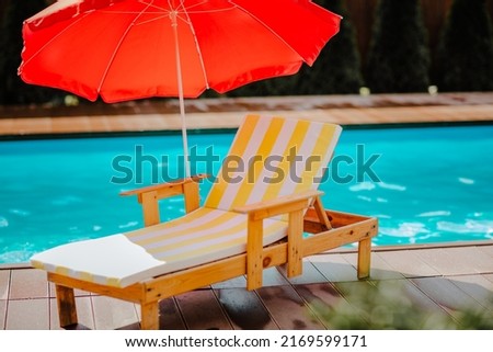 Stripped summer beach chair with red sun umbrella against blue swimming pool. Sunny day. No people Royalty-Free Stock Photo #2169599171