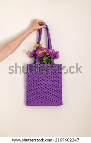 A woman's hand holds a purple crochet bag with purple flowers. Purple mesh bag with polyester cord on a white background. Zero waste concept.