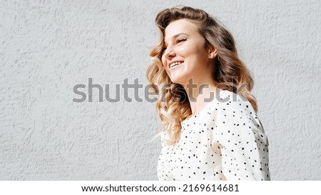 Laughing joyful young woman with curly hairstyle standing against gray wall and looking away, copy space. Portrait of stylish positive lady with toothy smile in white polka dot dress on sunny day.