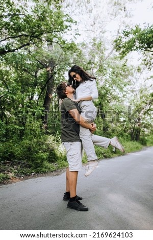 A bearded long-haired stylish man holds in his arms a brunette hippie woman in sunglasses gently hugs and smiles in nature in the park. Portrait, photo of the newlyweds.