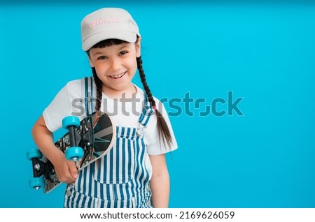 Funny child girl 6-7 years old with skate posing isolation blue background. concept children lifestyle, summer holidays, fashion. Layout copy space. Hold skateboard, children clothes cap, headdress,