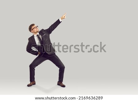 Funny businessman shows something isolated on copy space. Happy cheerful man in suit standing in funny pose isolated on light gray background and pointing index finger to right side. Business concept