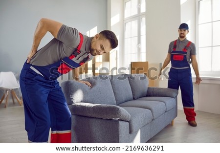 Worker feels sudden pain in his lower back when he tries to lift a heavy sofa. Two young men from a delivery service or a moving company can't carry a heavy couch because one of them gets injured Royalty-Free Stock Photo #2169636291