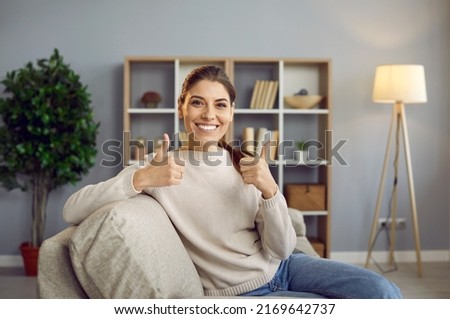 Cheerful satisfied beautiful young woman sitting on sofa in home interior, looking at camera, smiling and doing thumbs up gesture. Happy house owner or online course participant shows her satisfaction