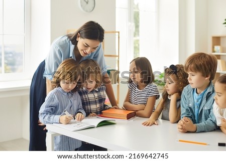 Young school teacher helping her happy elementary students who are writing in notebooks. Little children learning new things in class. Group of talented kids watching their classmate draw a picture