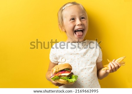 Cute little caucasian girl with blonde hair enjoying burger on a yellow background. Happy kid smiling and eating fast food burger