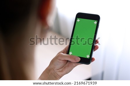 Chroma key mockup on smartphone in hand. Woman holds mobile phone iPhone and swipes photos or pictures left indoors of cozy home. Use green screen for copy space closeup
