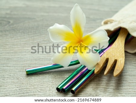 Reusable rainbow metal straws and bamboo fork in cotton carry bag with yellow and white plumeria blossom on light wood background, eco friendly lifestyle, reduce plastics and travel pono.